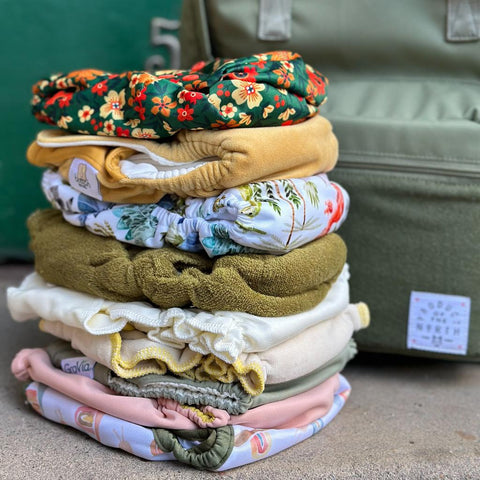 a cute stack of cloth diapers in muted neutral colors and prints alongside a green diaper bag in front of a green door