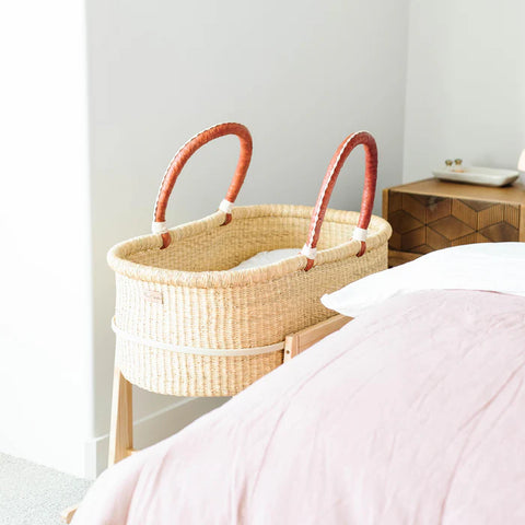 Woven baby Moses basket with red handles next to a bed in a bedroom setting.